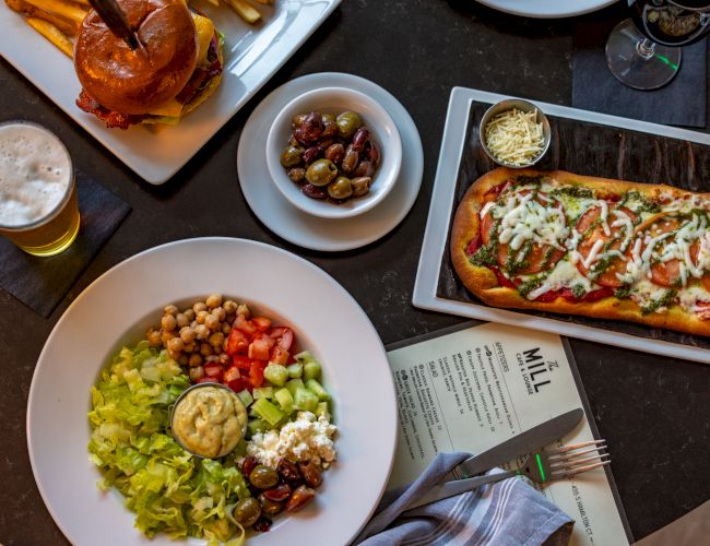 A colorful meal spread: a big salad bowl with greens, tomatoes, chickpeas, and toppings; a hot dog with toppings; a small bowl of olives; a plate with sliders and a beer; and a slice of bread with melted cheese, all on a dark table.