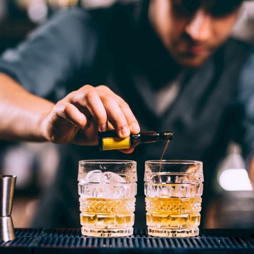 A person is making cocktails by adding a liquid to two glasses with ice on a bar counter, with a decanter and jiggers nearby.