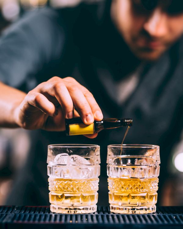 A person is making cocktails by adding a liquid to two glasses with ice on a bar counter, with a decanter and jiggers nearby.