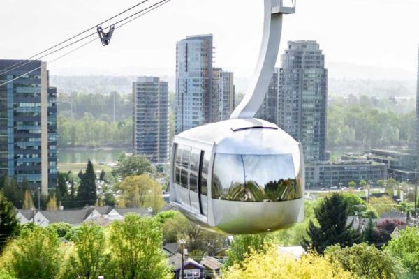 A modern gondola lift travels above a cityscape with tall buildings and lush greenery, against a backdrop of mountains and a cloudy sky.