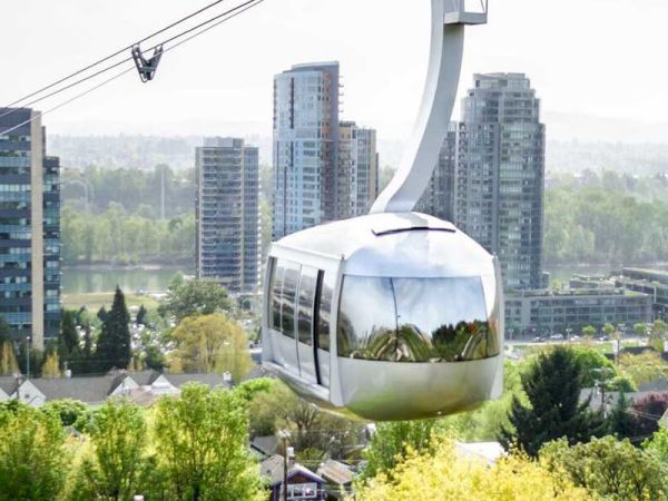 A modern gondola lift travels above a cityscape with tall buildings and lush greenery, against a backdrop of mountains and a cloudy sky.