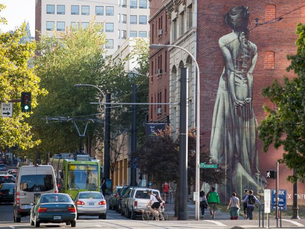 A bustling street scene with cars, a tram, and pedestrians. A large mural of a woman on a brick building adds artistic flair to the view.