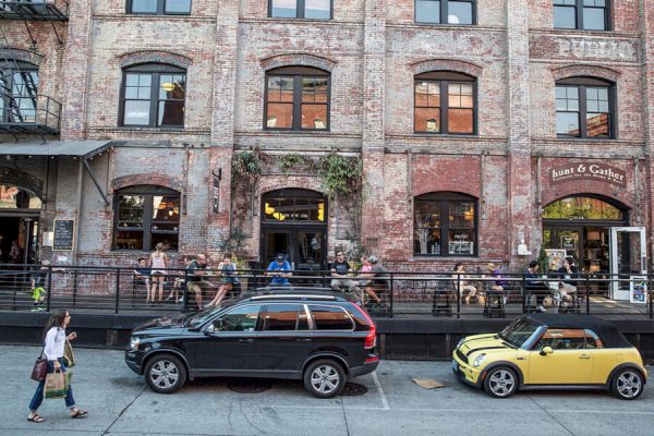 A street scene with parked cars in front of a brick building, people sitting outside a cafe or bar, and a pedestrian walking by.