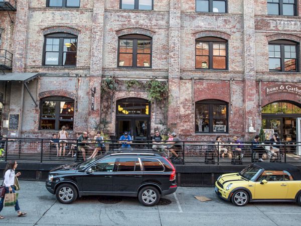 A street scene with parked cars in front of a brick building, people sitting outside a cafe or bar, and a pedestrian walking by.