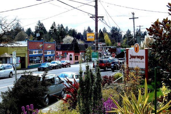 A small town street with parked cars, colorful buildings, a "Journeys" sign, and surrounding trees and greenery.