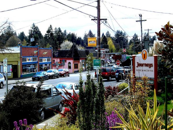 A small town street with parked cars, colorful buildings, a "Journeys" sign, and surrounding trees and greenery.