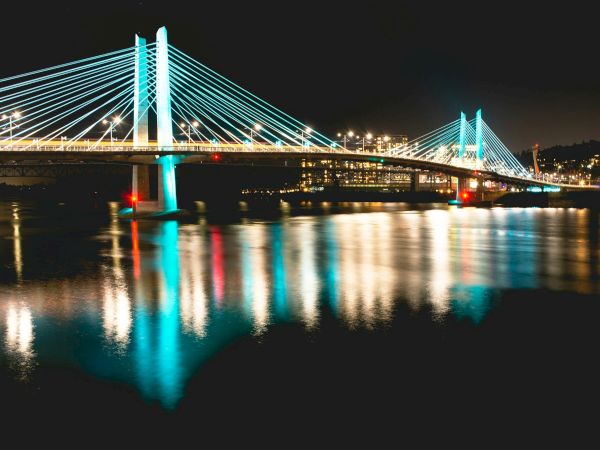A cable-stayed bridge lit up at night, reflecting beautifully on the calm river water below.