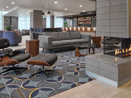 The image shows a modern lobby with a fireplace, stylish seating, a patterned rug, and a bar area in the background.