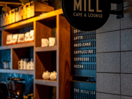 A cozy cafe sign reading “The MILL Café & Lounge” hangs by a wall; warm shelves with cups and drinks glow inside.