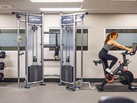A woman bikes at a stationary trainer in a gym, with weight machines and racks visible in the background, focused on her workout.