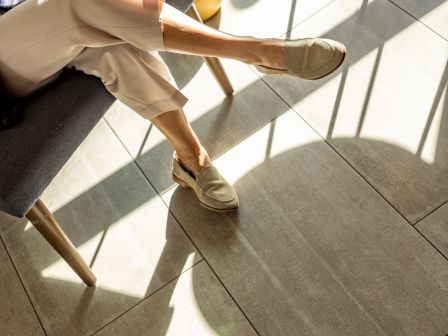 Someone sits on a chair with legs crossed, wearing beige pants and loafers, in a sunlit room with shadows and tiled floor, relaxing casually.