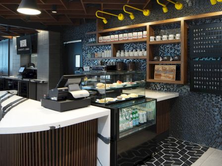 The image shows a modern café with a counter, display case of pastries, menu boards, shelves with jars, and tiled decor.