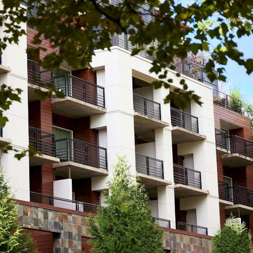 An apartment building with balconies is framed by green trees and a clear blue sky in the background.