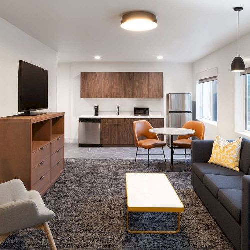 A modern living area with a kitchenette, TV, chairs, fridge, table, sofa, and decorative pillows, featuring a sleek and minimalist design.