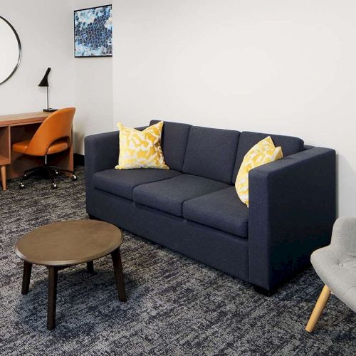 A room with a gray chair, blue sofa with yellow pillows, brown table, desk, orange chair, round mirror, and artwork on the wall.