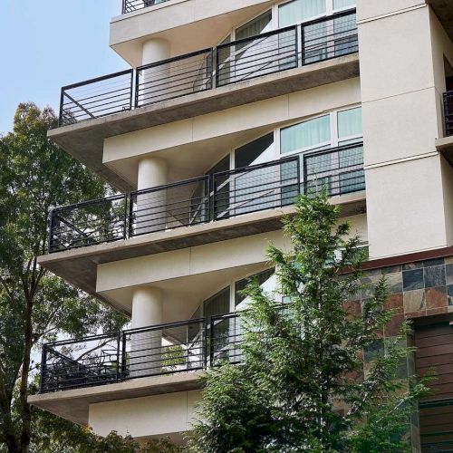 The image shows a modern building with multiple balconies and a surrounding of lush green trees.