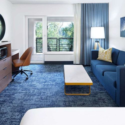 A modern hotel room features a TV, desk, sofa, and coffee table, with a balcony view and blue-themed decor and carpeting.