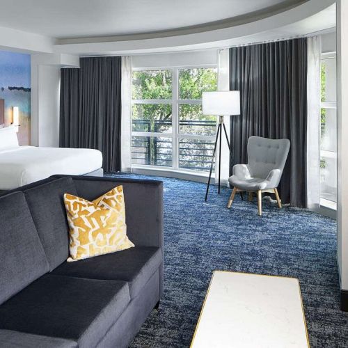 A modern hotel room with a living area, sofa with pillows, bed, large windows, chair, and lamp. Partial view of a bathroom included.