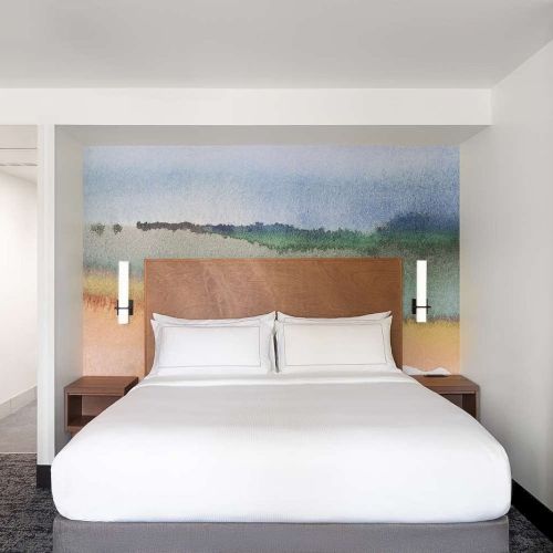 The image shows a neat hotel room with a double bed, white bedding, colorful headboard, and modern wall lights on each side.