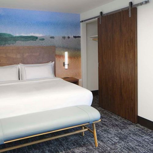 The image shows a modern hotel room with a bed, abstract wallpaper, sliding wooden door, bedside tables, and a view into a bathroom.