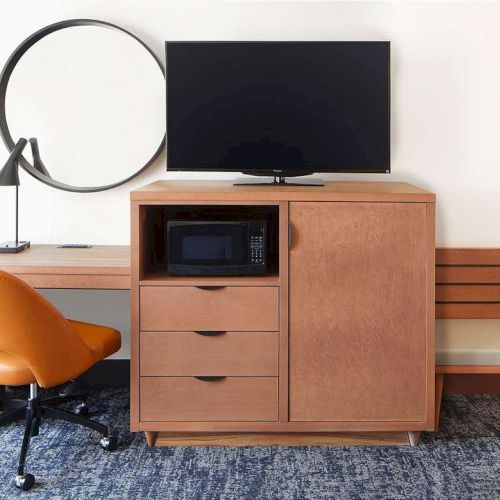 A room with a wooden desk, orange chair, lamp, large mirror, TV, microwave, and cabinet; set against a plain wall with a patterned carpet.