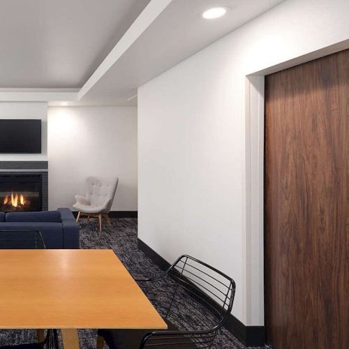 The image shows a modern living space with a fireplace, TV, blue sofa, wooden table, chairs, and a bedroom visible through a sliding door.