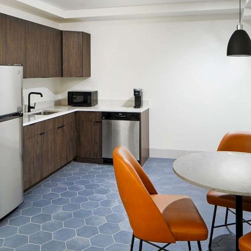 A kitchenette with wood cabinets, stainless steel fridge, microwave, coffee maker, sink, table, and leather chairs in a modern setting.