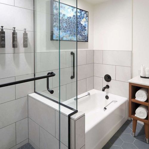 Modern bathroom with a glass shower, bathtub, tiled walls, towels on wooden shelves, a sink with a mirror, and some wall art.