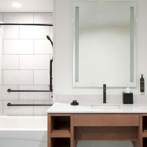 The image shows a modern bathroom with a tub, white shower curtain, vanity with a mirror, and wooden storage.