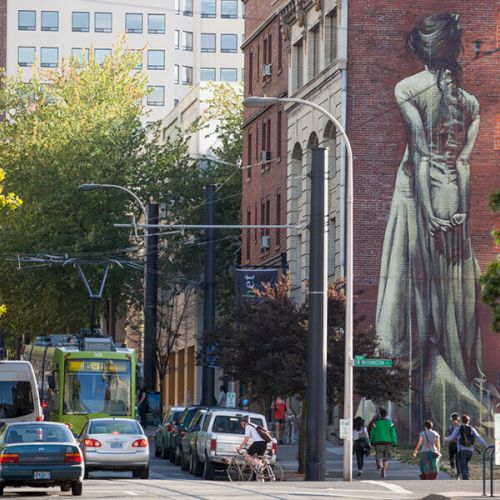A city street with cars and a tram, featuring a large mural of a woman in a dress on a brick building wall.