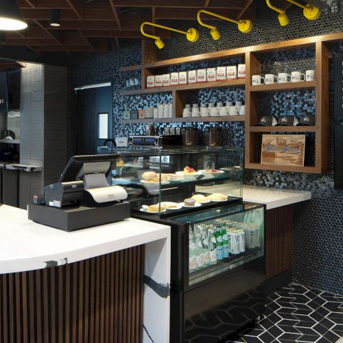 This image shows a modern café interior with a counter, menu board, pastries, drinks in a cooler, and decorative shelves to the side.