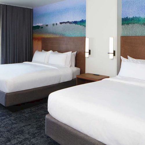 The image shows a hotel room with two large beds, a window with a view, modern decor, and a colorful mural on the wall.