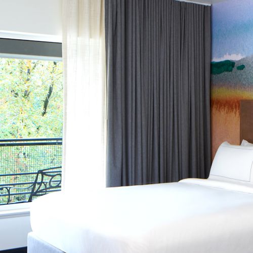 A cozy room with a neatly made bed, large window, curtains, and a colorful mural on the wall, overlooking a leafy outdoor view.