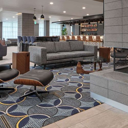 A modern lounge with a cozy fireplace, stylish seating, and a patterned carpet, next to a bar area with stools and a variety of drinks displayed.