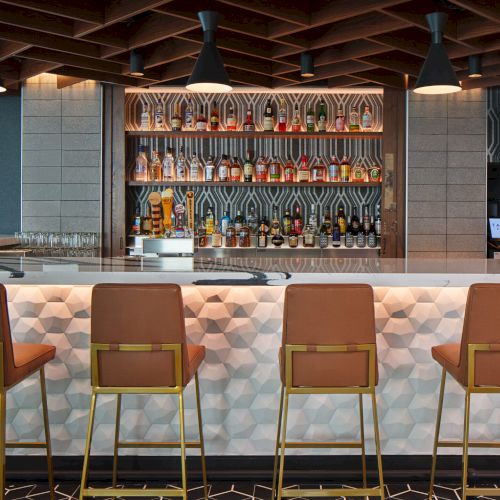 The image shows a stylish bar with leather chairs, a well-stocked shelf of bottles, and modern lighting.