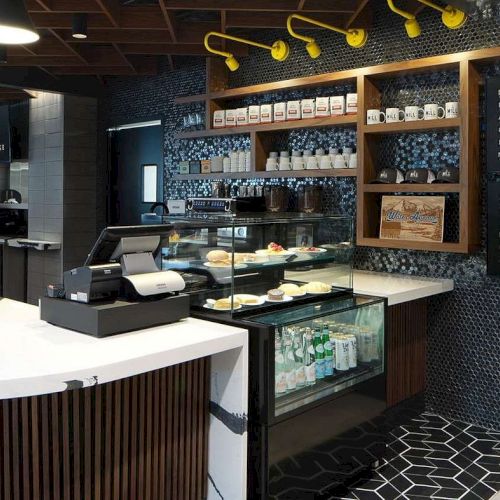 Modern café interior with a menu board, display of pastries, drinks, and shelves with jars, surrounded by stylish lighting.