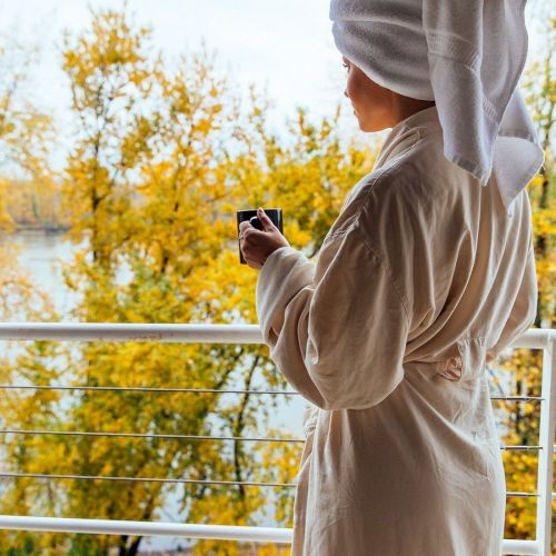 A person in a robe with a towel on their head holds a cup, enjoying a view of trees with yellow leaves from a balcony.