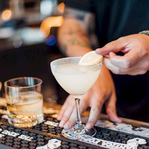A person is garnishing a cocktail with a lemon slice; a bottle and a glass are nearby on the bar.