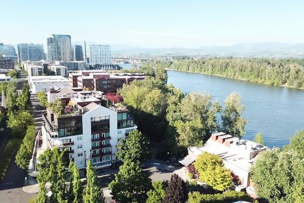 An aerial view of a riverside cityscape with modern buildings, lush greenery, and a river under a clear blue sky.