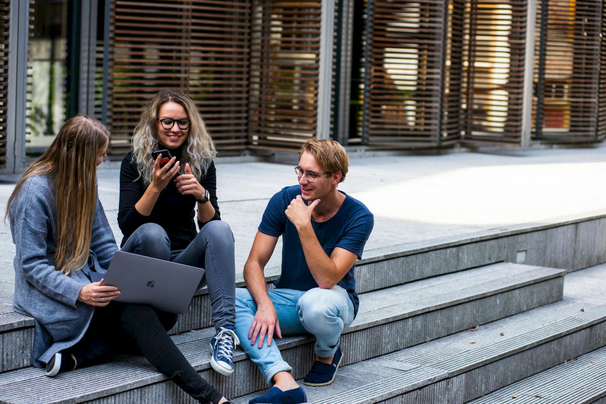 Three people are sitting on steps outside, engaged in conversation. Two have laptops, and one is holding a smartphone.