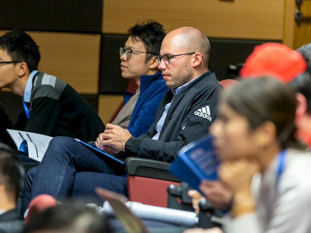 People are attentively seated, possibly at a conference or lecture, holding papers and notebooks, focused on the speaker.