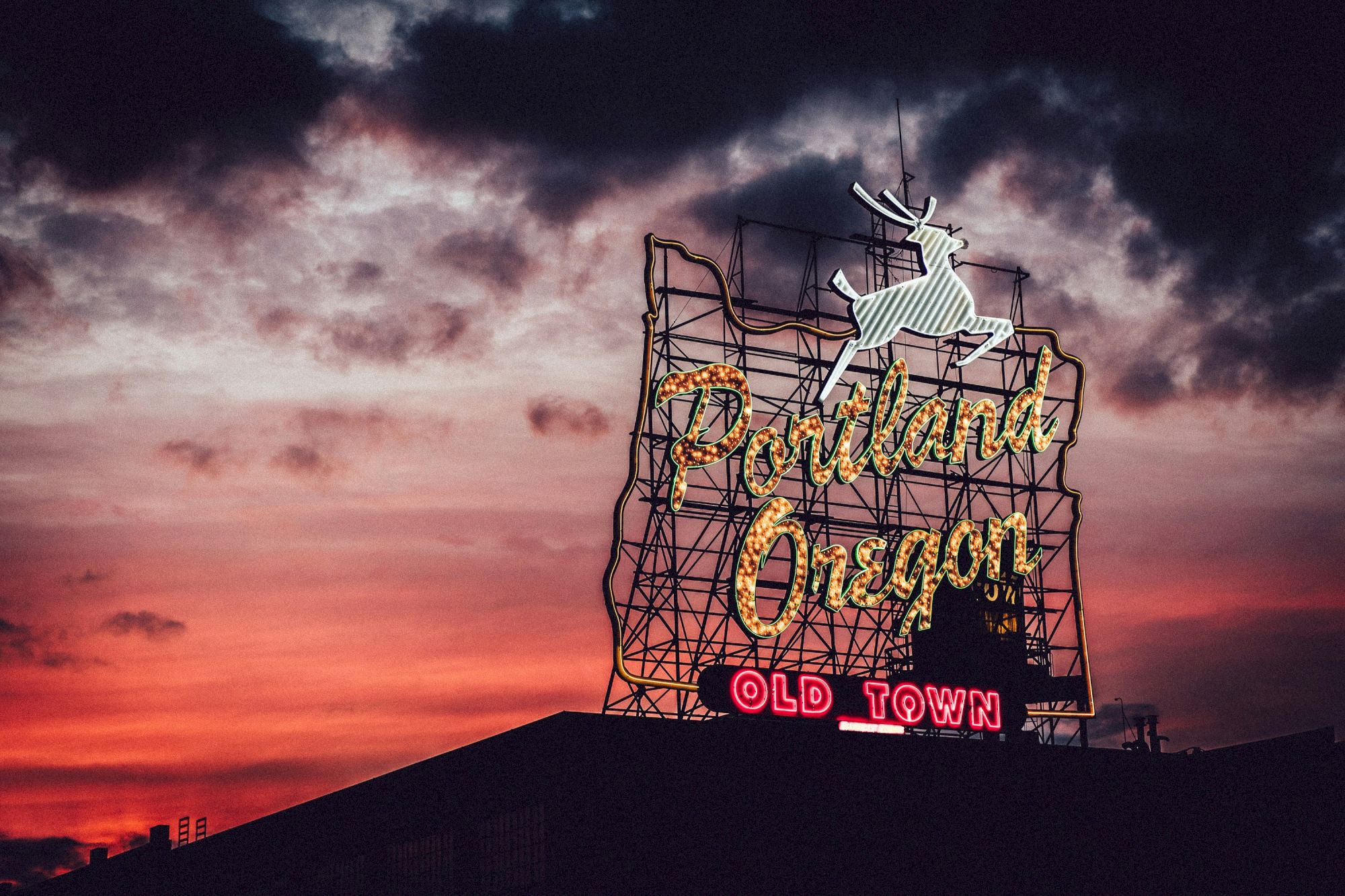 A neon sign reads "Portland Oregon Old Town" at sunset, with a leaping deer above, set against a dramatic cloudy sky.