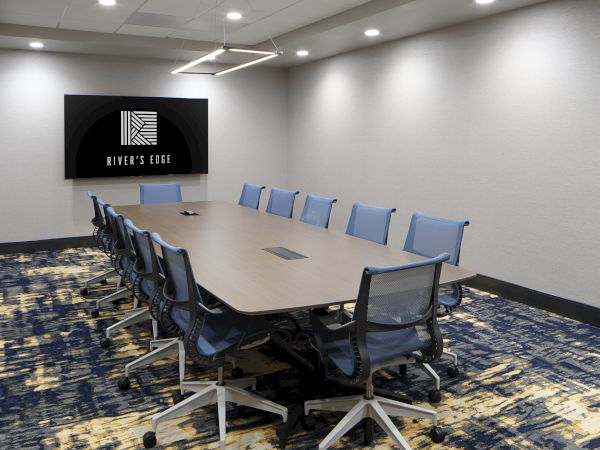 The image shows a modern conference room with a large table, several chairs, a screen on the wall, and a patterned carpeted floor.