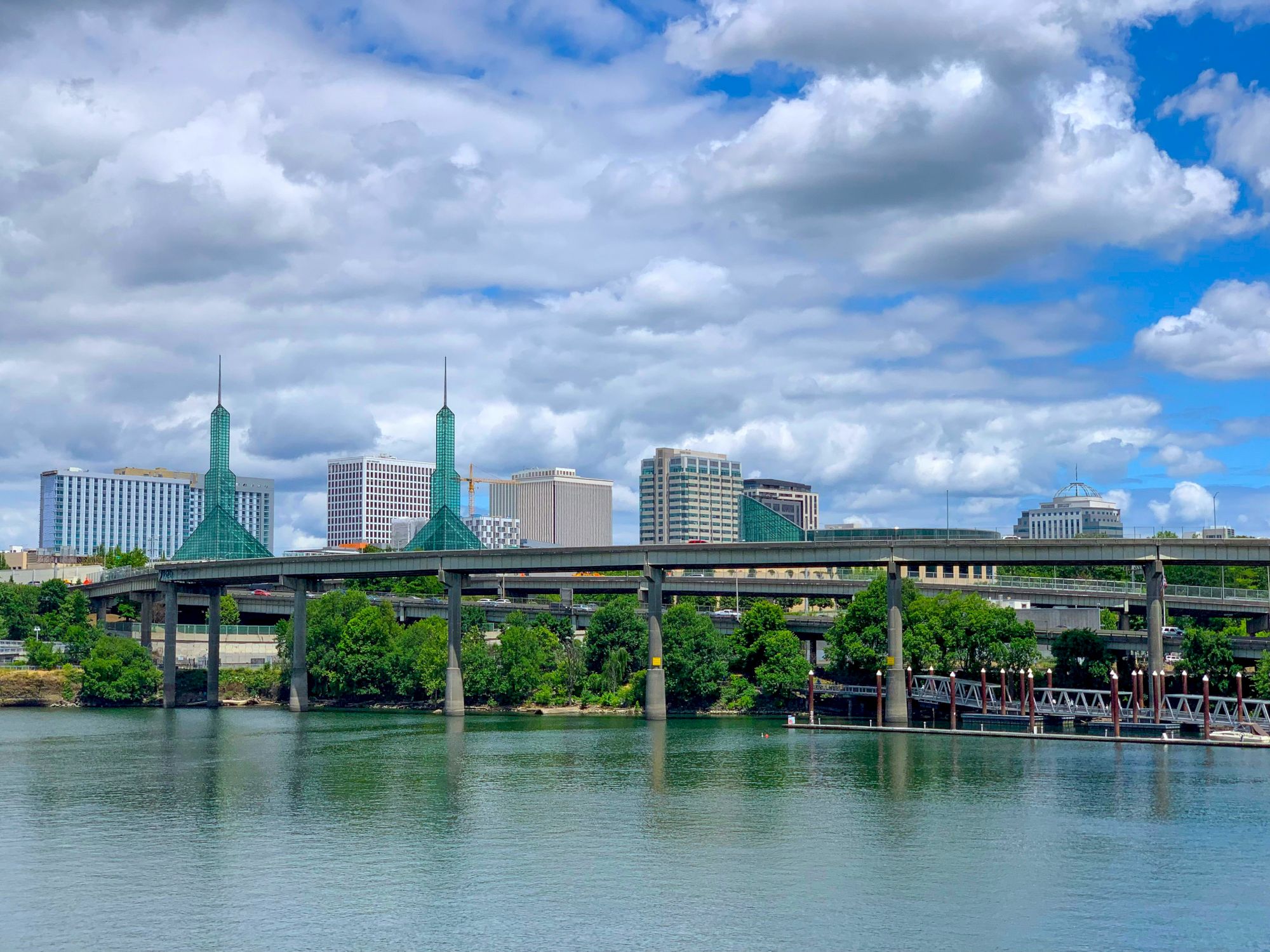 A waterfront view with a highway bridge, modern buildings, and trees beneath a cloudy blue sky is shown in the image.