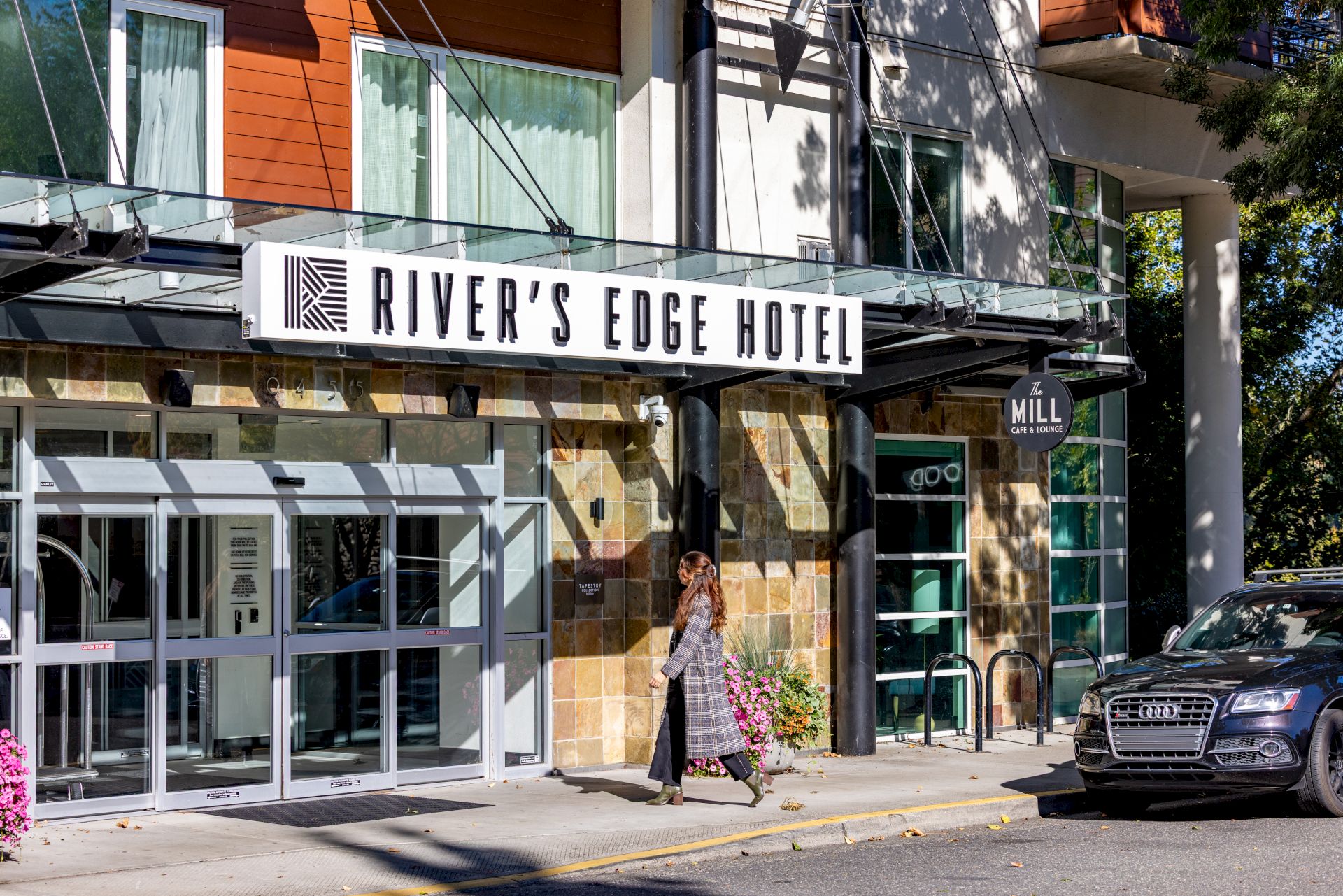 A street view of River&rsquo;s Edge Hotel with a glass storefront, a couple walking by, and a parked car by the entrance.