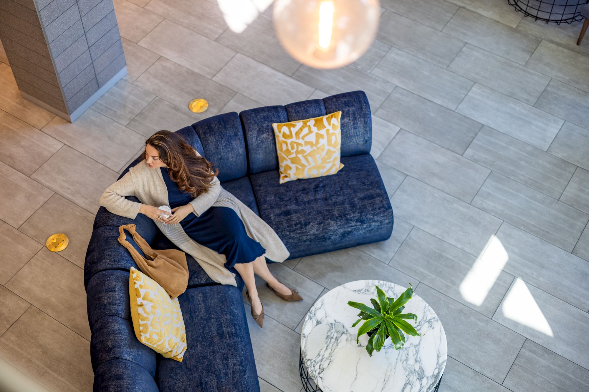 Aerial living room: a blue sectional with yellow cushions surrounds a round marble coffee table; a woman lounges on the couch, plant on table, bright sunlight streaming in.