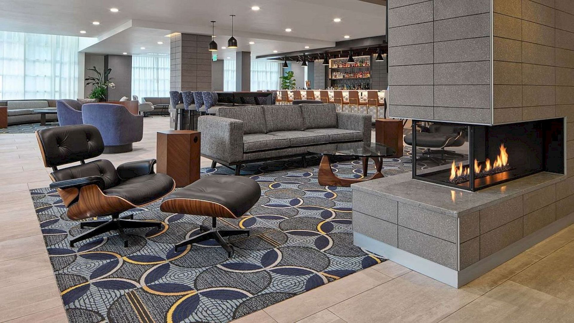 A modern hotel lobby with a cozy lounge, fireplace, contemporary seating, geometric patterned carpet, and a bar area in the background.