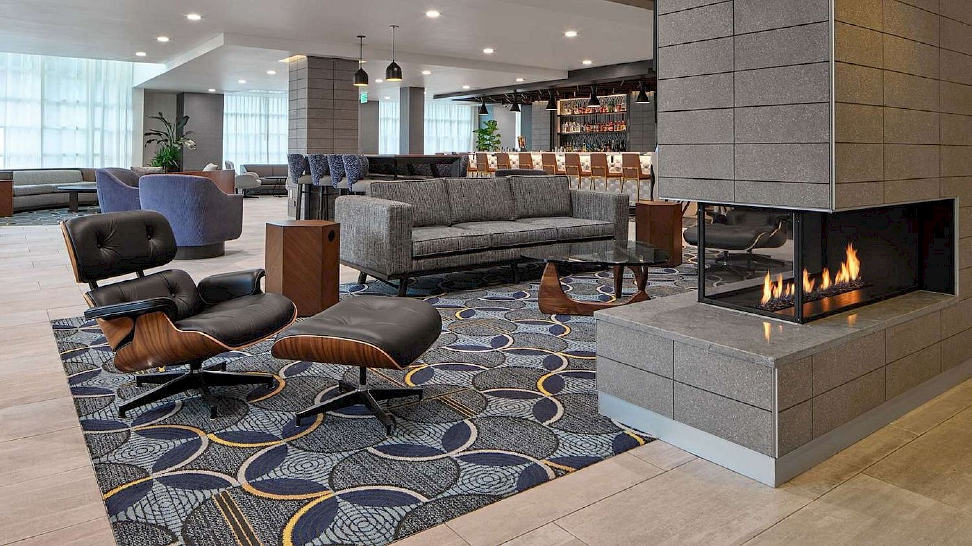 A modern hotel lobby with a cozy lounge, fireplace, contemporary seating, geometric patterned carpet, and a bar area in the background.