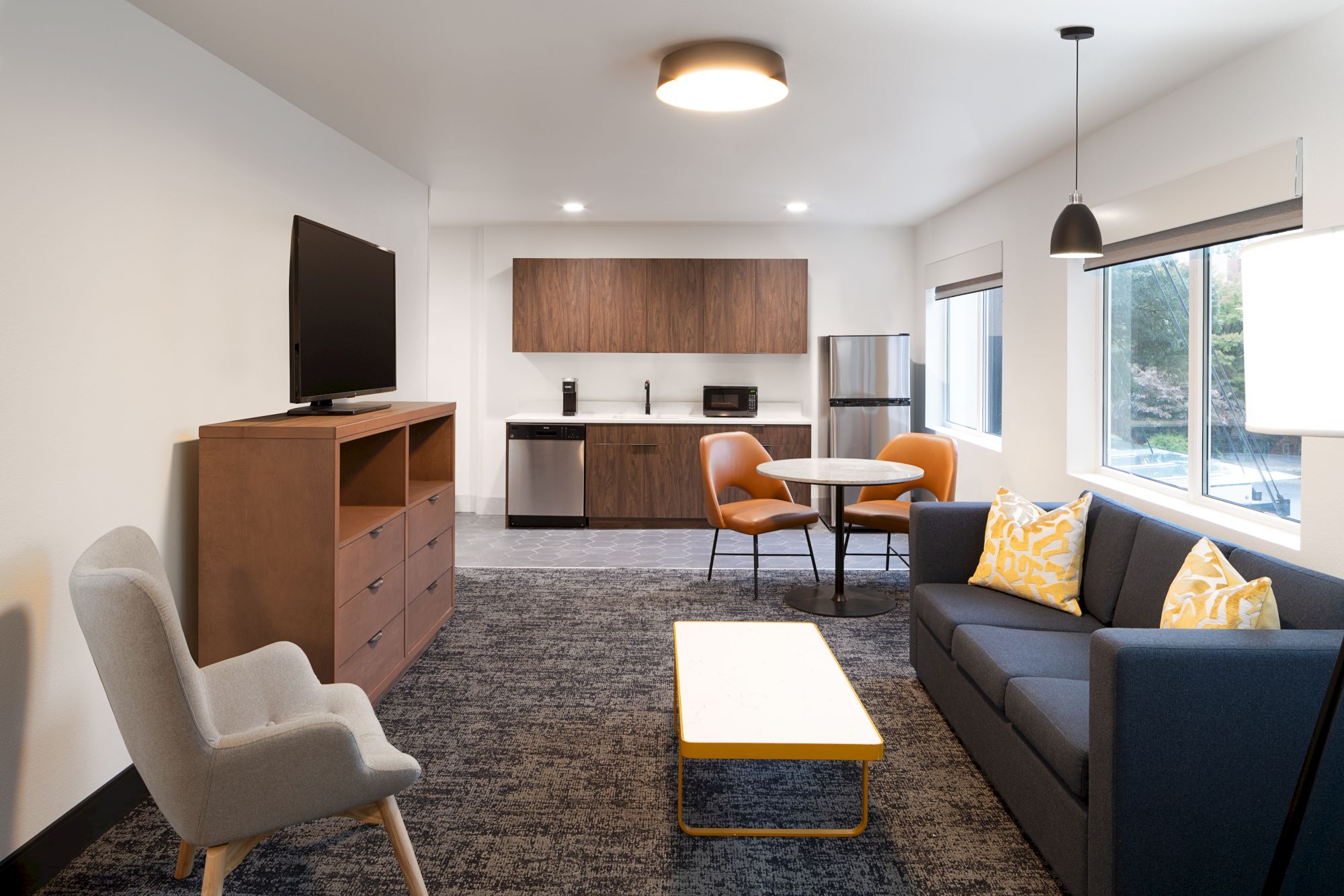 A modern, compact living area with a sofa, armchairs, coffee table, and a small kitchenette; bright, with a half-wall of windows.