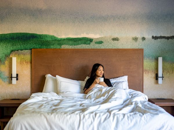A woman sits up in a large bed with white sheets, in a room with a painted landscape mural and two wall sconces on either side of the headboard.